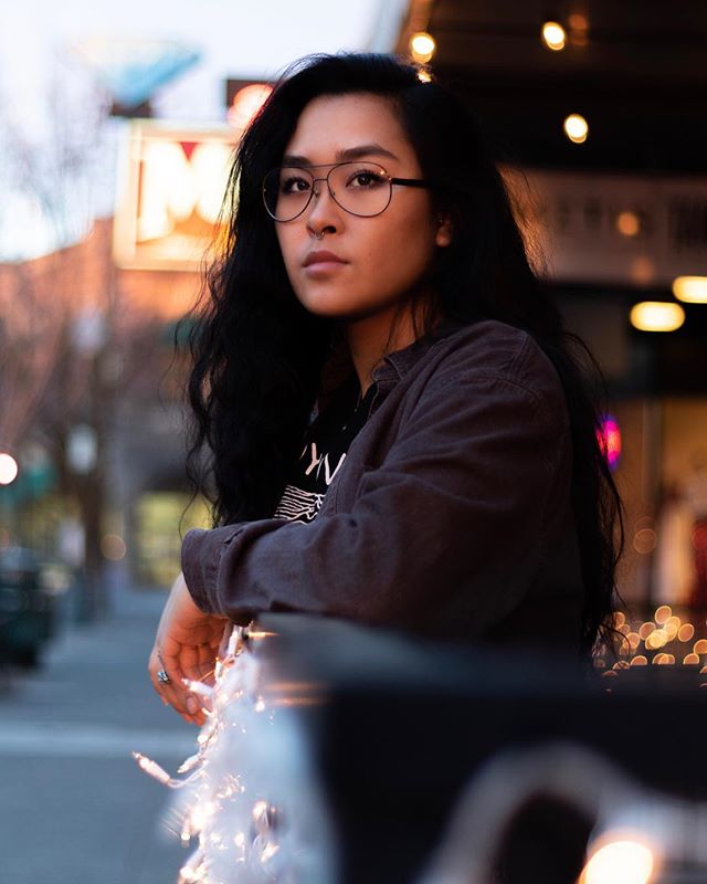 A young woman with glasses poses in an urban setting at night.