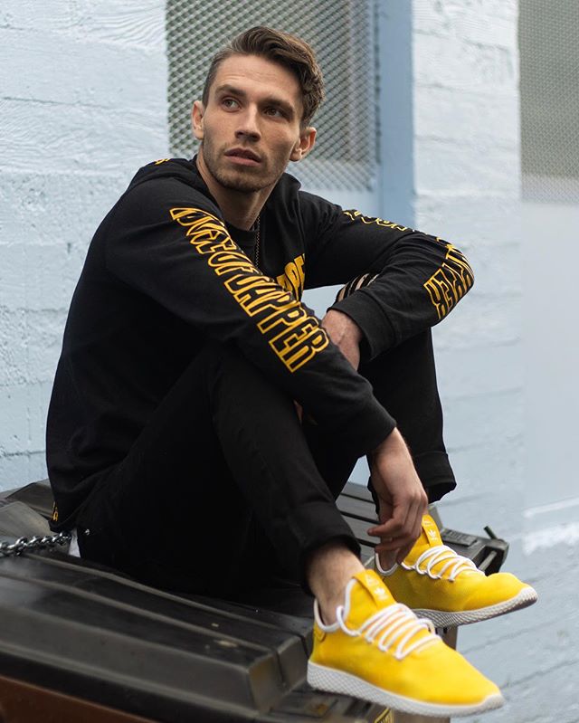 A man sitting in an urban setting, wearing black clothing and yellow Adidas sneakers.