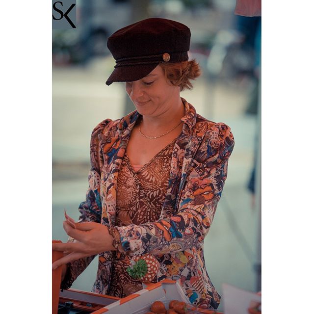 A woman in a patterned jacket and hat smiles at a market stand.