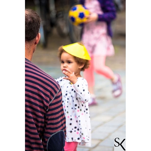 A toddler with a yellow hat stands next to an adult on a city street, with a child holding a ball in the background.