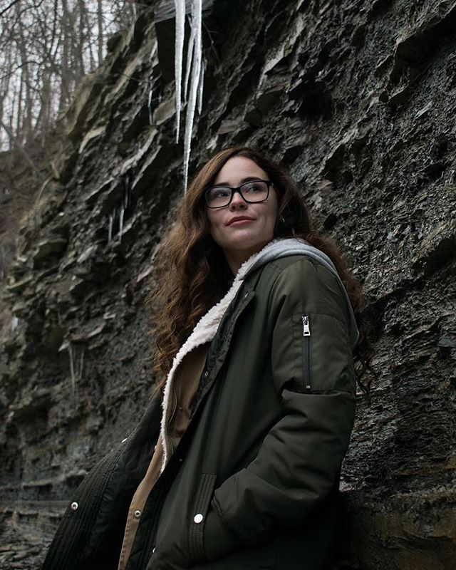 A woman in glasses and a green jacket stands near a rocky cliff in a natural outdoor setting.