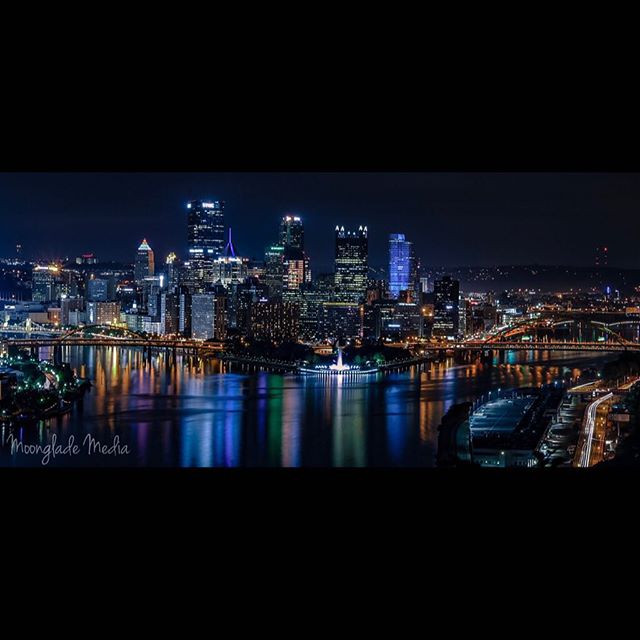 Nighttime cityscape featuring a river reflecting city lights, bridges, and skyscrapers, capturing an urban waterfront scene.