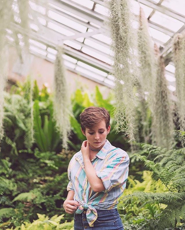 A young woman stands in a lush greenhouse, surrounded by greenery, wearing a pastel shirt and denim jeans.