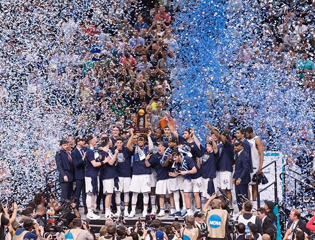 A basketball team celebrates winning a championship with confetti and a trophy, cheered on by a large crowd.