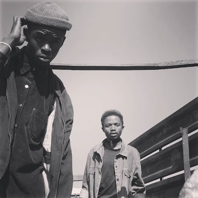 Two stylish young men pose in a monochrome outdoor shot, exuding serious intensity.