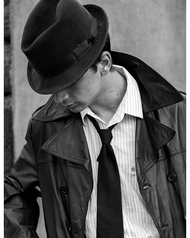 A man in a fedora and trench coat looks down in a dramatic, vintage-style black and white image.