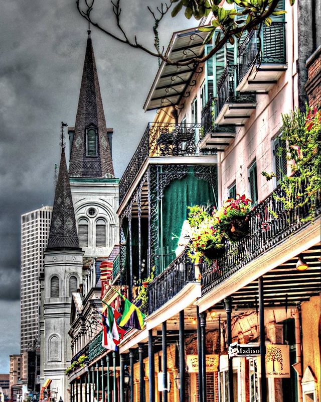 A vibrant street view of New Orleans with historic architecture, flags, and ornate balconies under a cloudy sky.