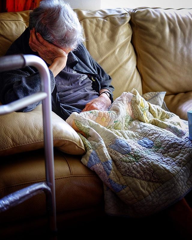 An elderly woman rests on a couch with a quilt, near a walker, portraying a scene of quiet reflection and aging.