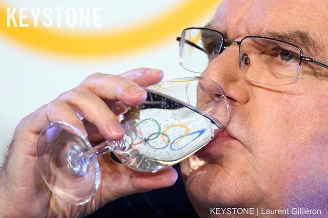 A man drinks from a glass of water, Olympic rings are visible through the glass.