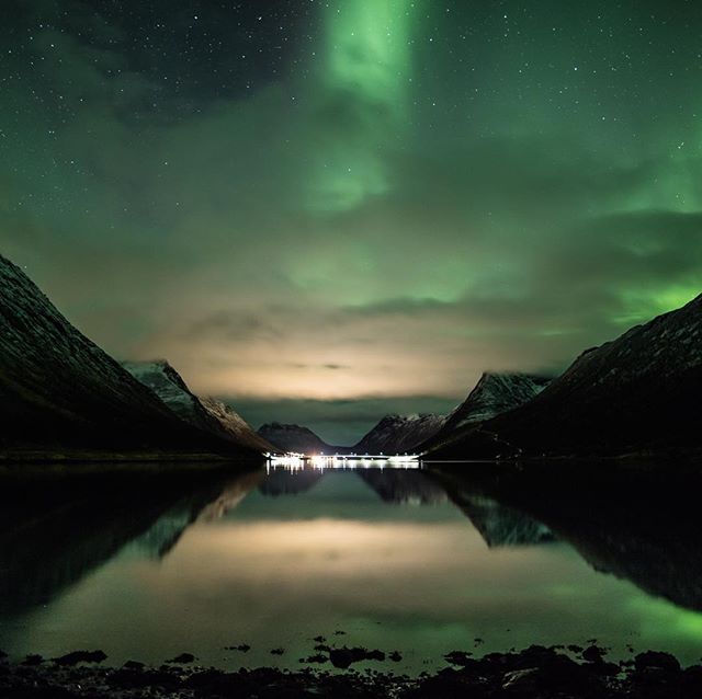 A serene night landscape featuring the aurora borealis reflected in a tranquil lake surrounded by mountains.