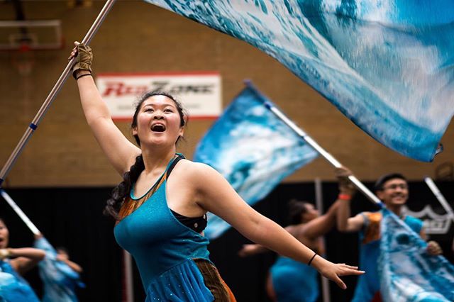 A color guard performs indoors, waving large blue and white flags with energy and precision.