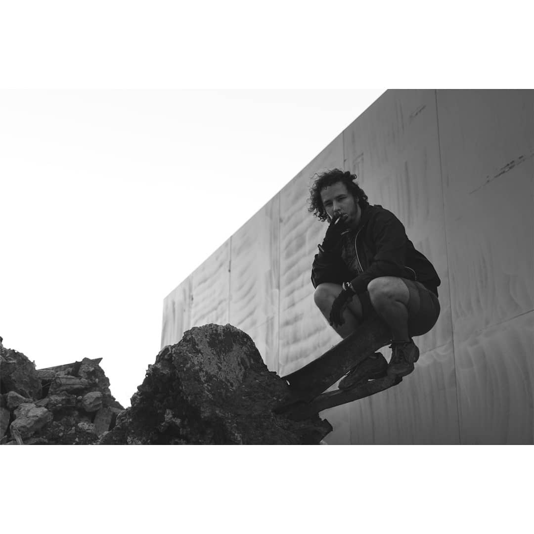 A man with curly hair smokes a cigarette while squatting on a rock against an industrial wall in a black and white outdoor scene.