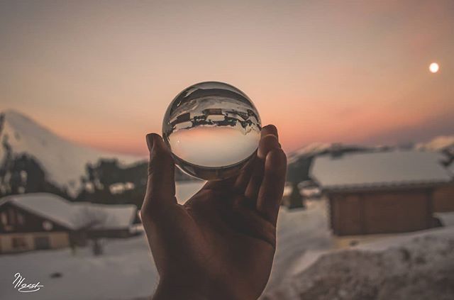 Hand holds a crystal ball reflecting a winter landscape with snowy mountains and a sunset sky.