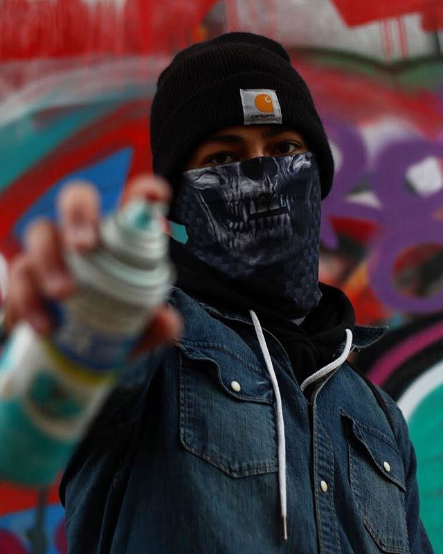 A graffiti artist with a skull mask holds a spray can in front of colorful graffiti art.