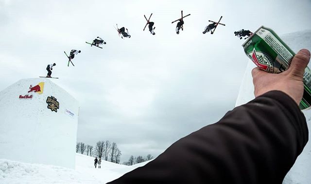 A skier performs a trick off a snow jump as a hand holds a beer can in the foreground.