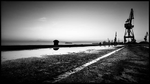 Black and white image featuring cranes at a dock by the sea, evoking a sense of industrial activity and maritime transport.