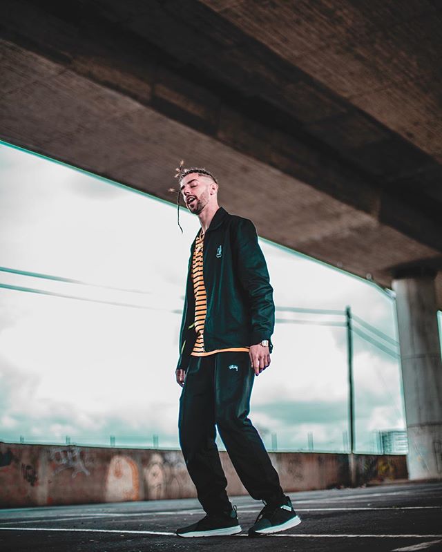 Man in streetwear walks confidently under a bridge, blending urban style with a relaxed attitude.