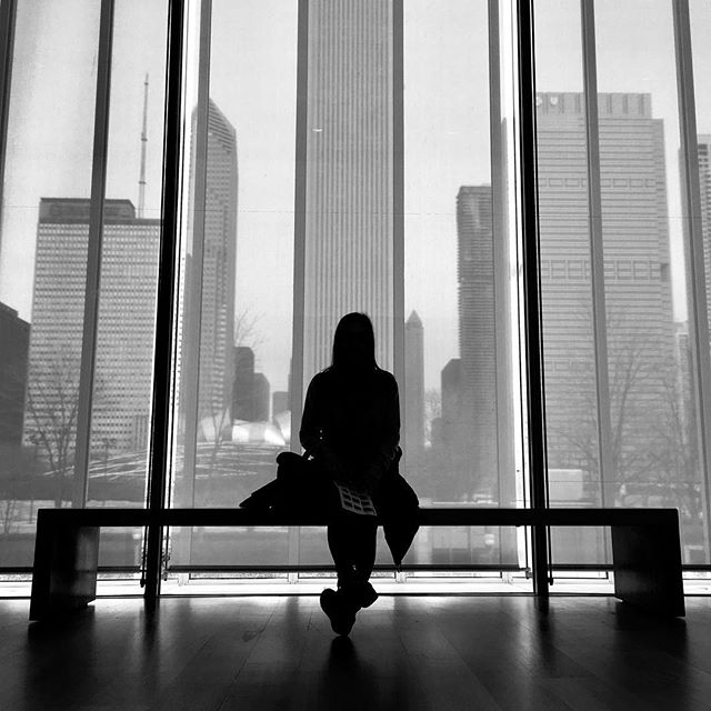 A silhouetted woman sits on a bench, gazing out at the city skyline through a large window.