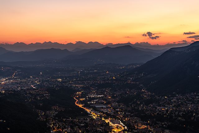 A serene cityscape at night with city lights nestled between mountains and an orange sky at sunset.