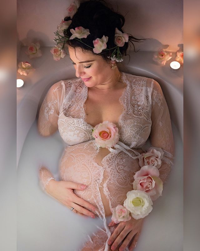 A pregnant woman bathes in a milk bath with flowers, wearing a lace robe and flower crown.