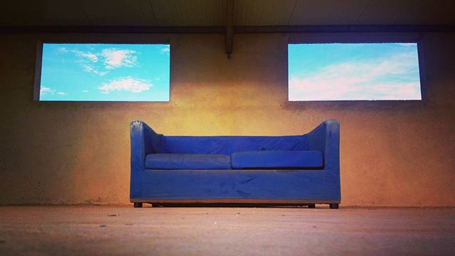 A blue sofa sits beneath two windows showing a serene blue sky with clouds in a minimalist setting.