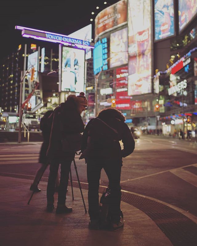 People with camera on a street at night with brightly lit billboards in an urban setting.