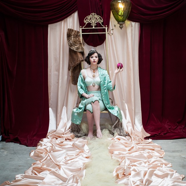 A woman wearing a tiara and robe sits, holding a pink ball in a vintage styled scene.
