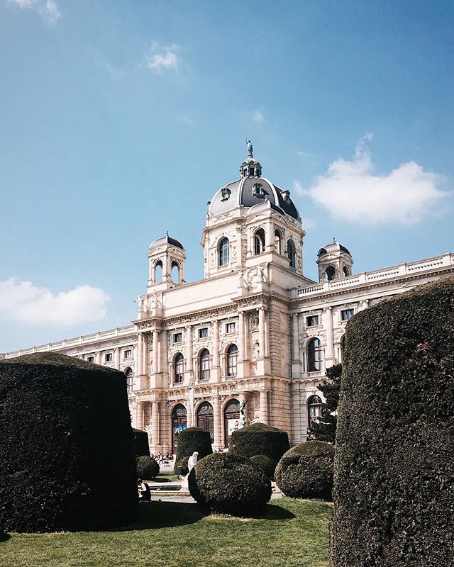 A grand building with a dome surrounded by green bushes under a blue sky in a city.