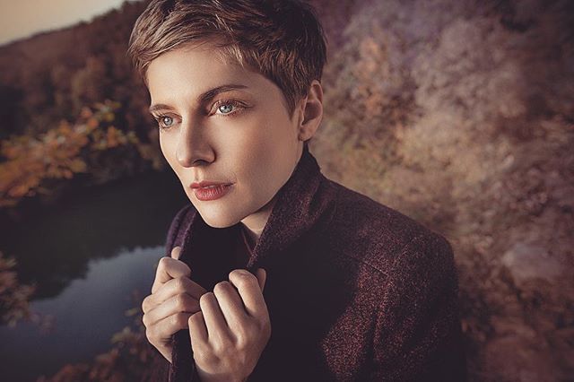 A portrait of a woman with short hair in a coat, posing outdoors in a natural setting.