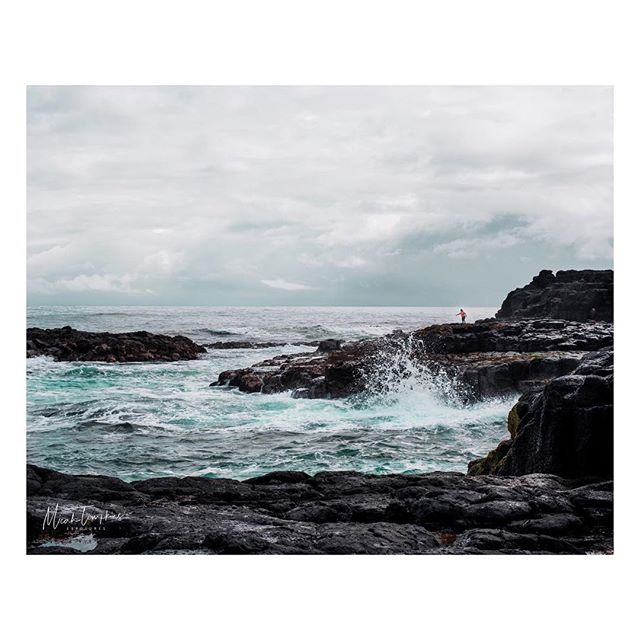 A person stands on rocks near the ocean as waves crash against the shore on a cloudy day.