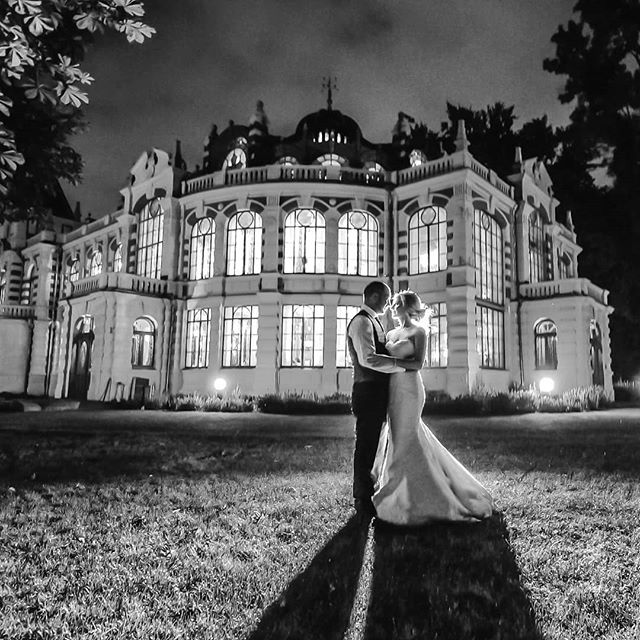 A bride and groom embrace in front of a brightly lit mansion at night.