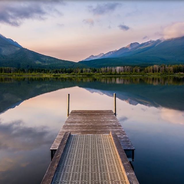 A tranquil lake reflects mountains with a wooden pier extending into the calm water.