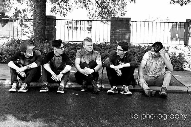A black and white image features five young men sitting together outdoors in a casual, urban setting.