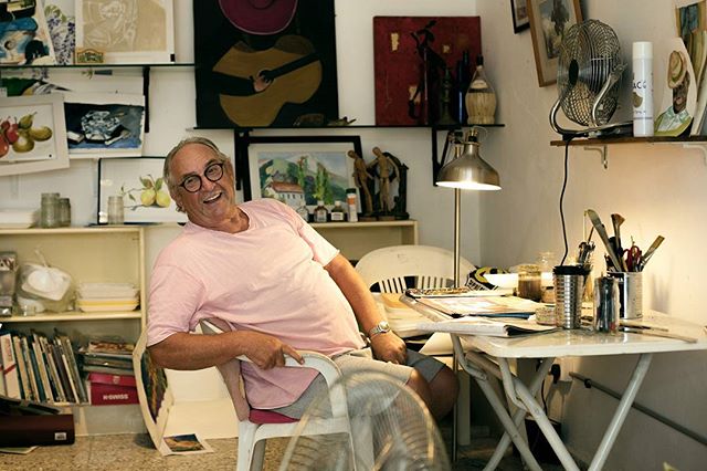 An artist smiles in their studio, surrounded by artwork and supplies.