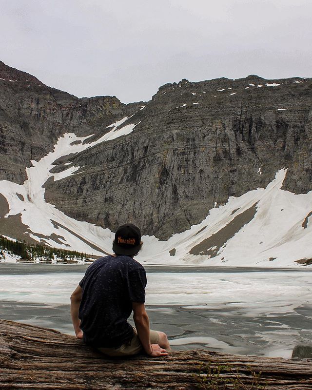 A man sits on a log, gazing at an icy lake and rugged mountains under a cloudy sky.