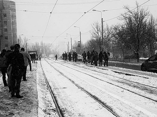 A black and white image shows people clearing snow from tram tracks on a city street during a winter snowfall.