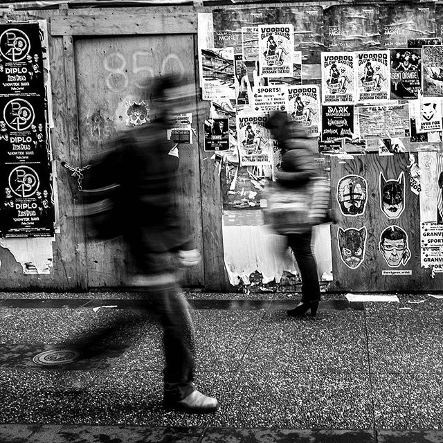 Black and white street scene with blurred figures walking past a wall covered in posters and street art.