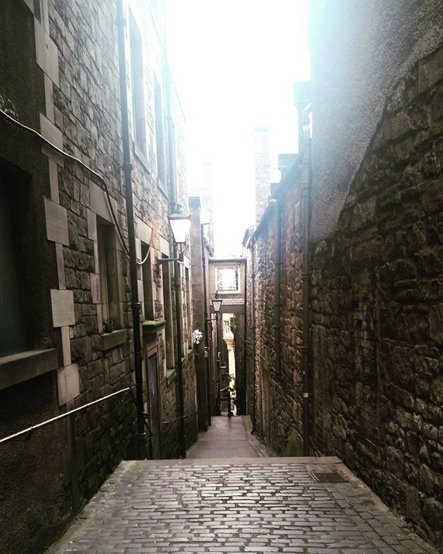 A narrow cobblestone street in a historic European city with stone buildings on either side and a bright light at the end.