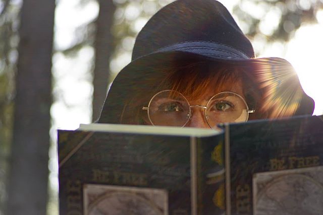A woman with glasses and a hat is reading a book in a forest setting with a shallow depth of field.