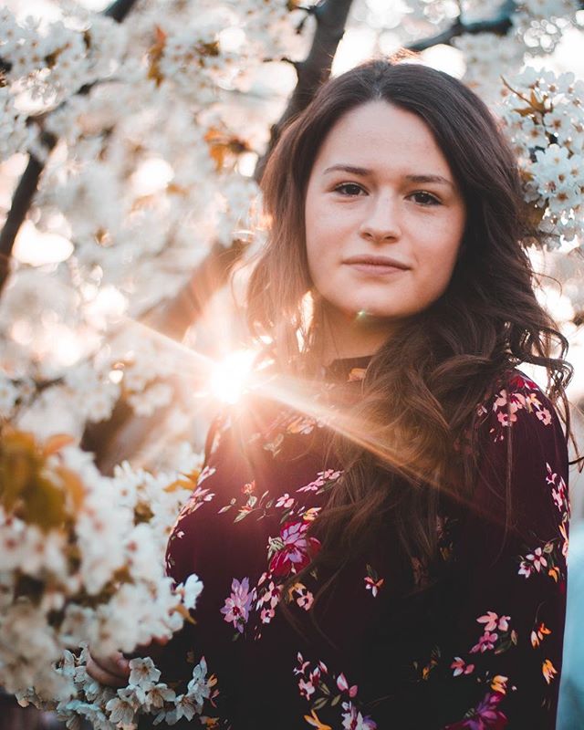 A woman in a floral dress stands among white blossoms in soft light, creating a serene and natural portrait.