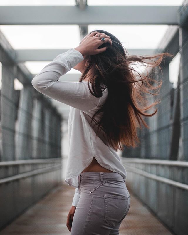 A young woman with long brown hair stands on a bridge, her hand on her head, in a stylish candid shot.