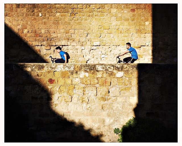 Two men cycle on a brick wall in a sunlit urban setting, promoting a fit and active lifestyle.