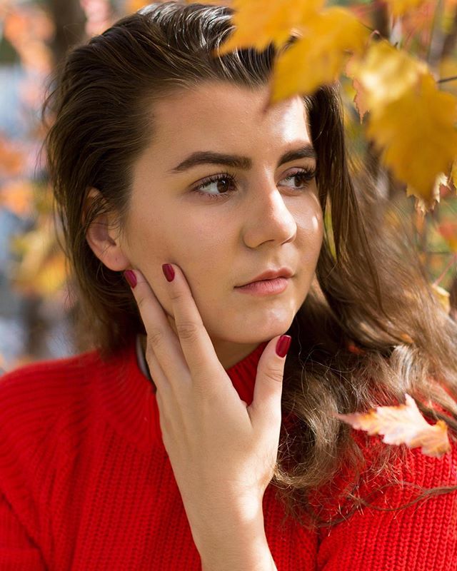 A young woman in a red sweater looks thoughtfully off camera amidst autumn leaves.