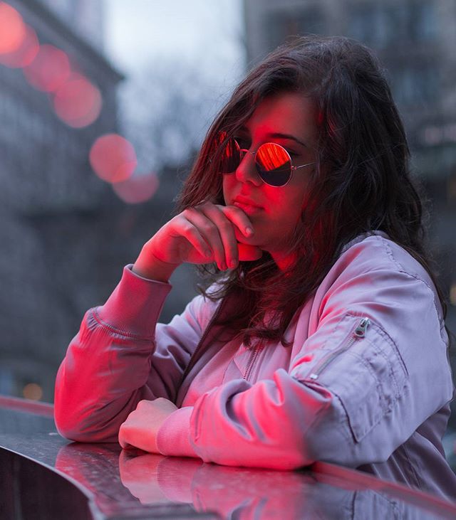 A stylish woman in sunglasses and a jacket contemplates in an urban setting, highlighted by a red glow.