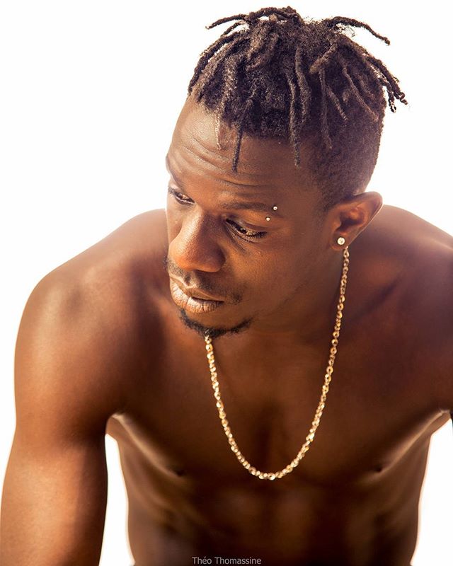 Intense portrait of a shirtless man with dreadlocks and a gold chain, posing against a white background.