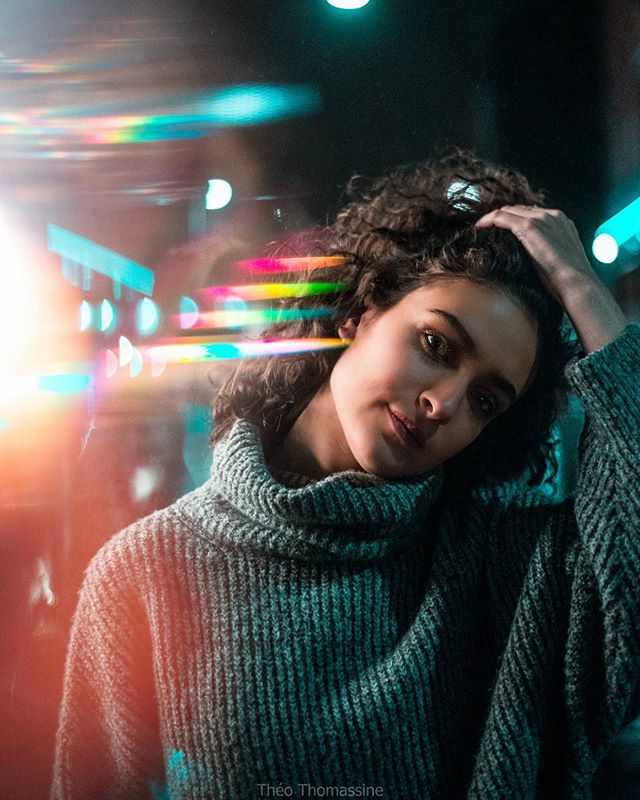 A woman with curly hair poses in a cozy turtleneck sweater amidst vibrant city lights.