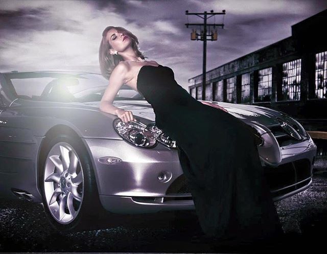 A woman in a black dress poses elegantly on a silver car in an urban setting at night.