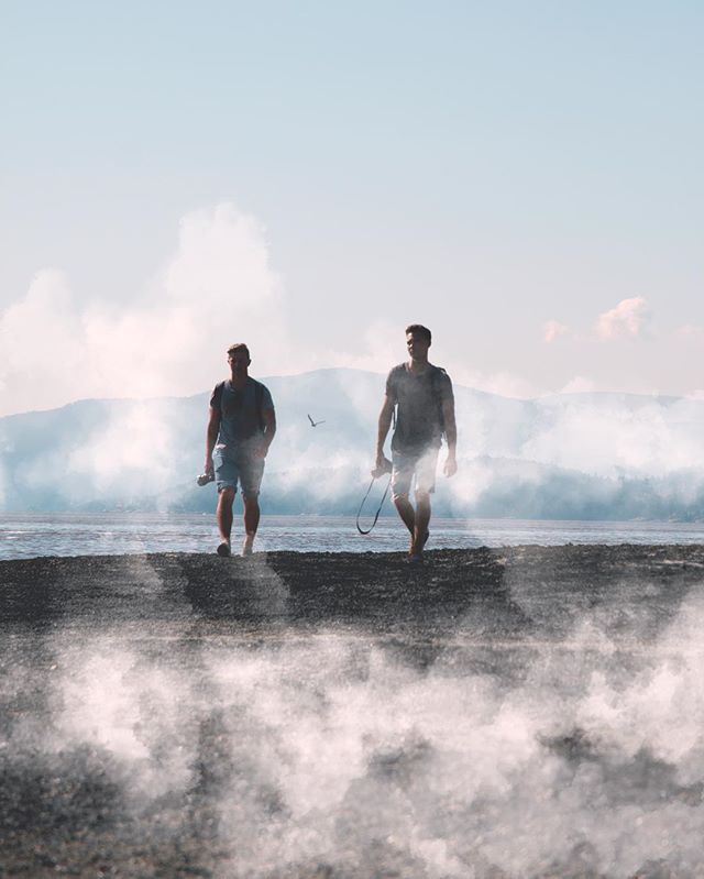 Two men walk along a misty beach on a bright day near a mountain range.