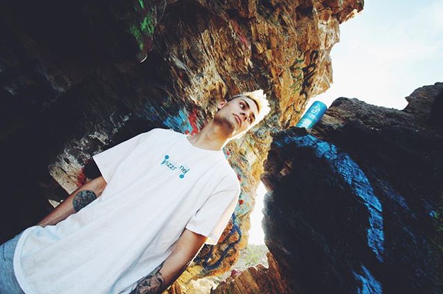 A blond man poses in a rocky cave environment, lit by natural light for an editorial style shot.
