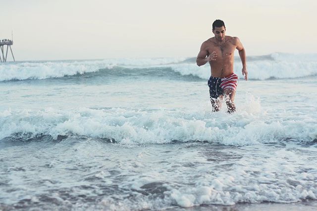 Athletic man running through ocean waves wearing patriotic swim trunks at the beach.